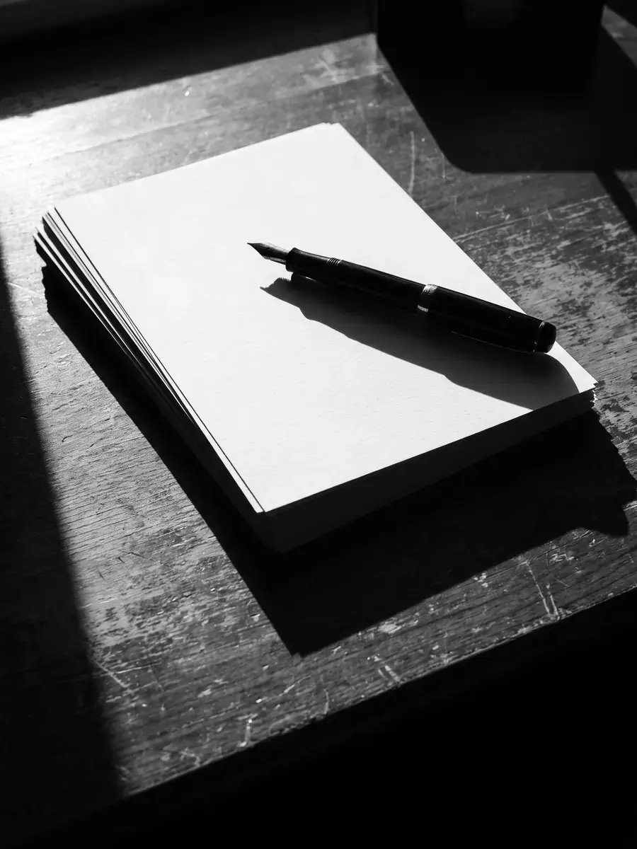 A stack of blank paper and a pen on a bare desk, black and white